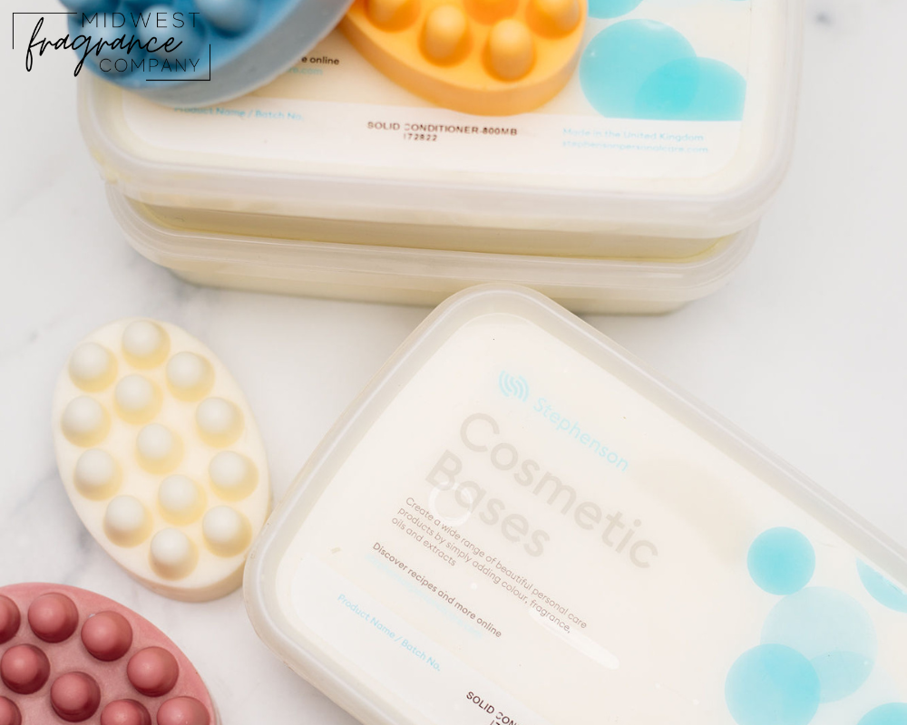 Close-up of Stephenson Cosmetic Bases solid conditioner packaging, surrounded by colorful massage bar soaps. The Midwest Fragrance Company logo is visible in the top left corner. The image showcases high-quality solid conditioner bases and handcrafted massage bars on a clean, marble surface.
