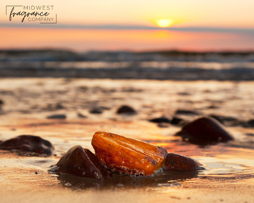 A caramel colored piece of sea glass nestled among rocks on a sunset shoreline depicting a fragrance called Brazilian Crush Cheirosa 71, a dupe from Sol de Janeiro.