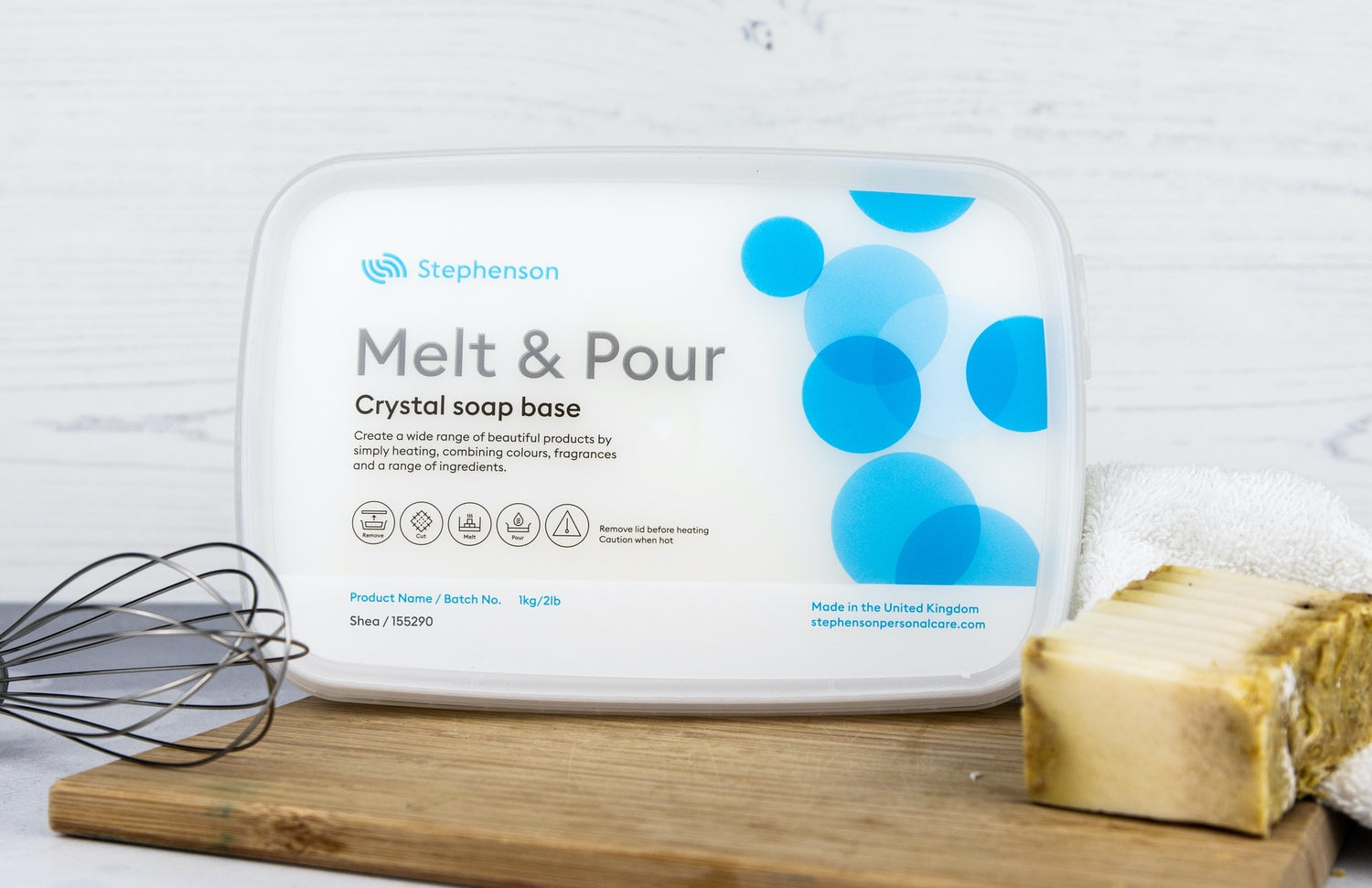 A blue and white 2 lb. tub of soap base on a wood cutting board with a bar of soap and a wire whisk on a white background.