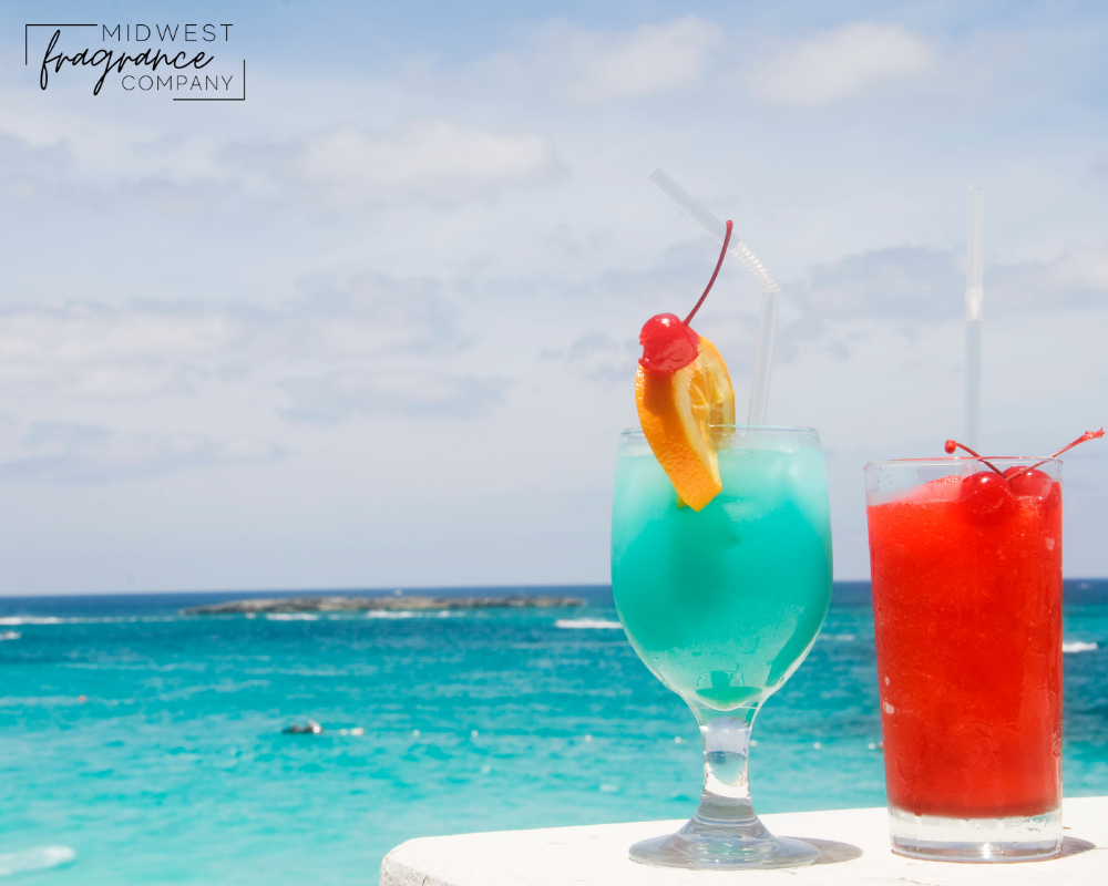 Glasses of tropical drinks in blue and red on a white table in front of the ocean depicting a fragrance called Jamaica Me Crazy.