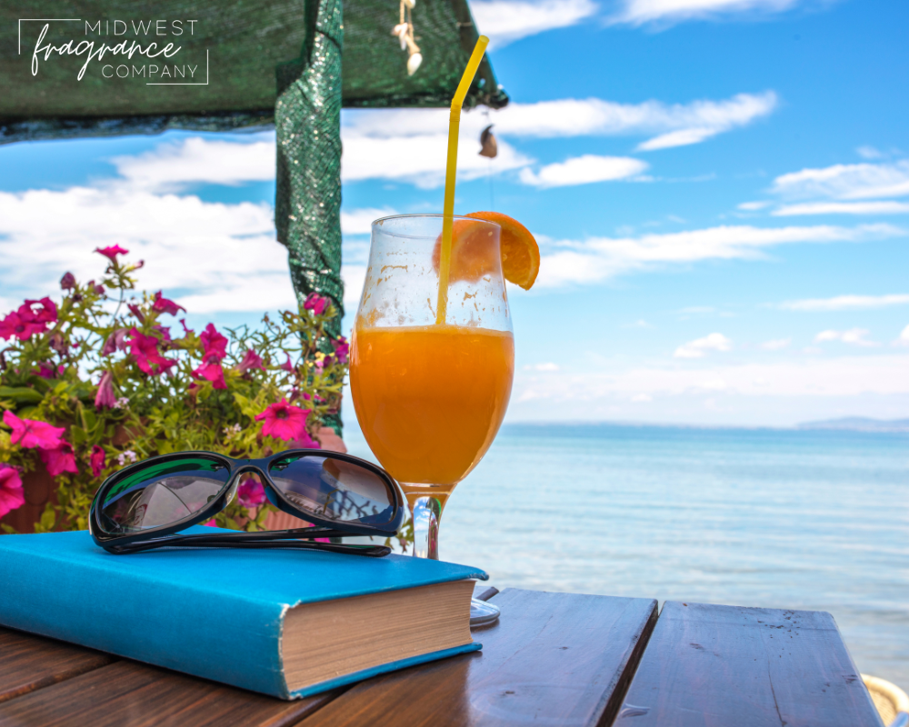 A tropical drink and straw on a wooden table with a book, sunglasses, and flowers in front of the ocean illustrating a fragrance called Endless Weekend.