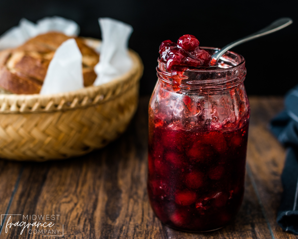 A mason jar of cranberry apple marmalade with a spoon and loaf of fresh baked bread in the background on a rustic table.
