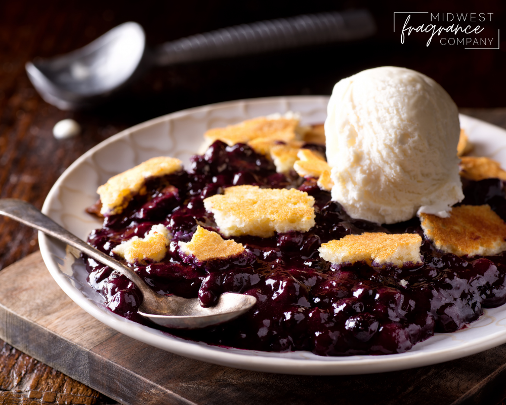 A white ceramic bowl with blueberry cobbler and vanilla ice cream on a wood cutting board and ice cream scoop in the background.